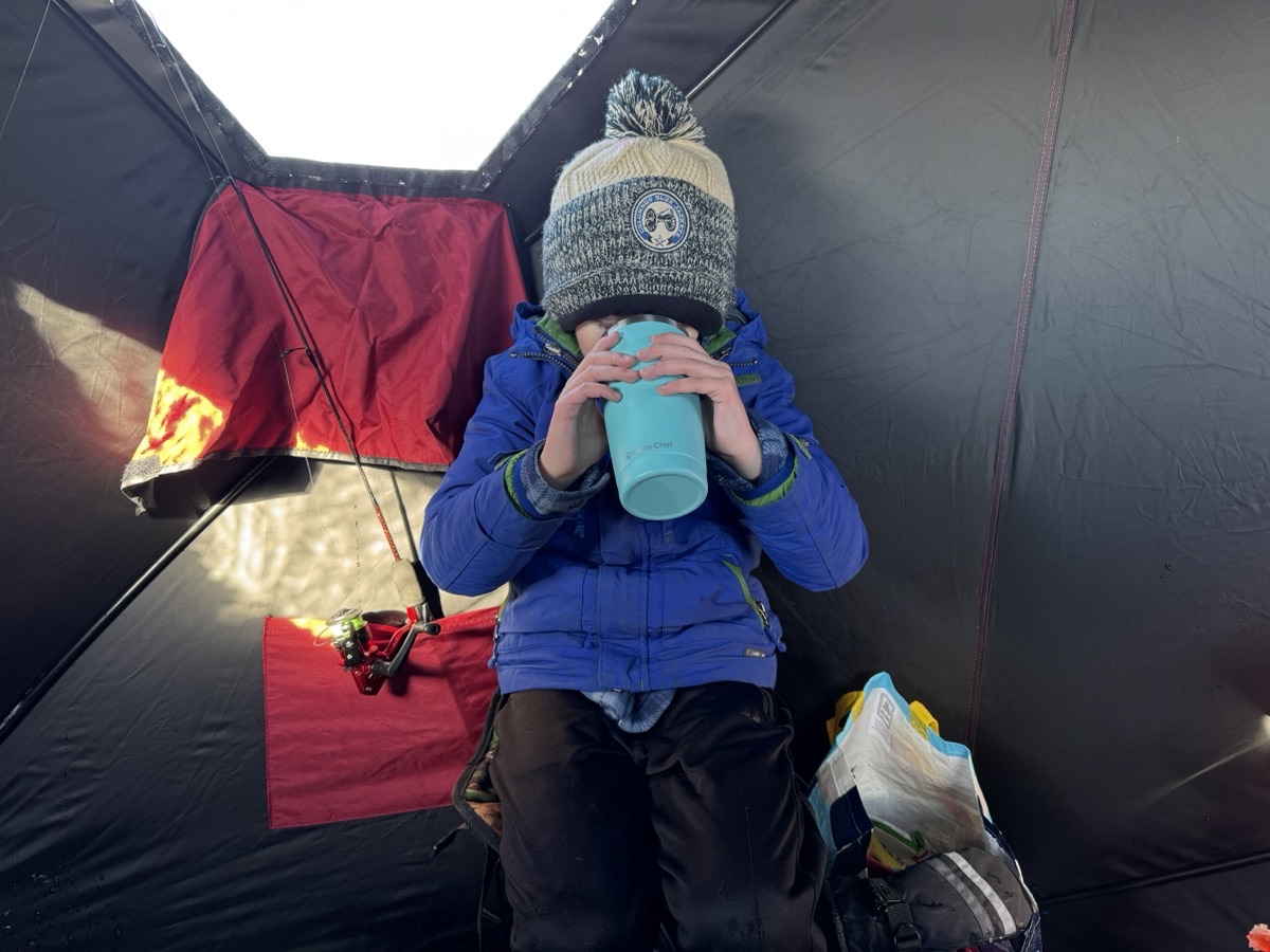 Julian drinking hot cocoa from a tumbler in the ice fishing shelter